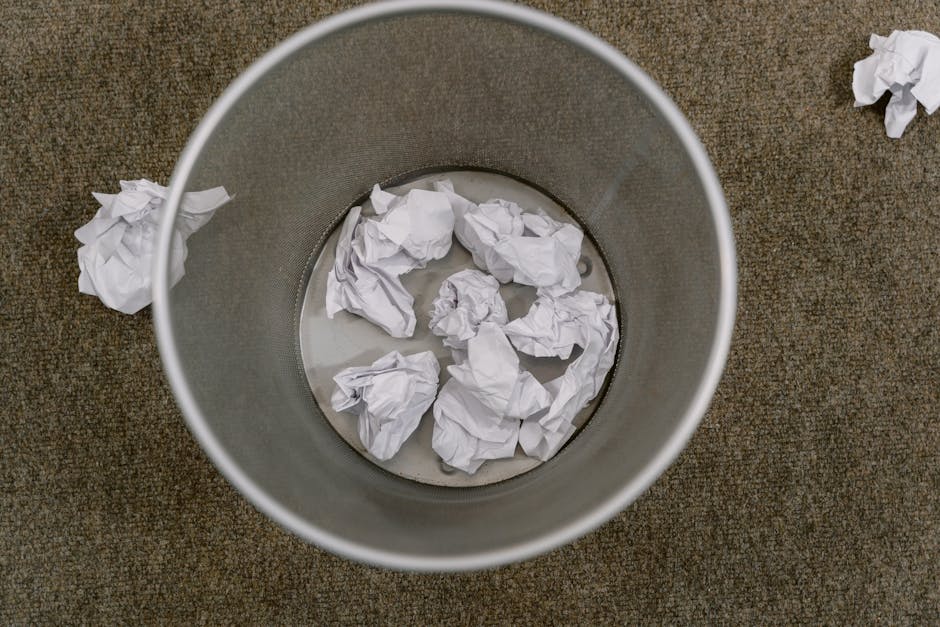 An overhead view of a cylindrical metal rubbish bin placed on a brown, textured carpeted surface. Inside the bin, several crumpled white paper sheets are partially submerged in a small amount of clear
